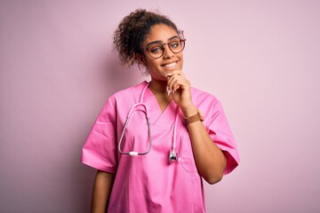 Poster - African american nurse girl wearing medical uniform and stethoscope over pink background looking confident at the camera with smile with crossed arms and hand raised on chin. Thinking positive.