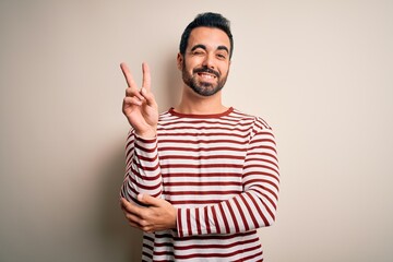 Canvas Print - Young handsome man with beard wearing casual striped t-shirt standing over white background smiling with happy face winking at the camera doing victory sign. Number two.