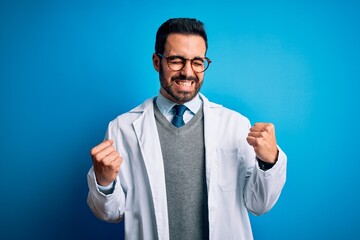 Sticker - Young handsome doctor man with beard wearing coat and glasses over blue background excited for success with arms raised and eyes closed celebrating victory smiling. Winner concept.