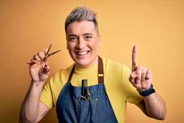 Young modern professional hairdresser man holding scissors over isolated yellow background surprised with an idea or question pointing finger with happy face, number one