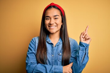 Poster - Young beautiful asian woman wearing casual denim shirt and diadem over yellow background with a big smile on face, pointing with hand and finger to the side looking at the camera.