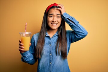 Canvas Print - Young beautiful asian woman drinking healthy glass of orange juice over yellow background stressed with hand on head, shocked with shame and surprise face, angry and frustrated. Fear and upset