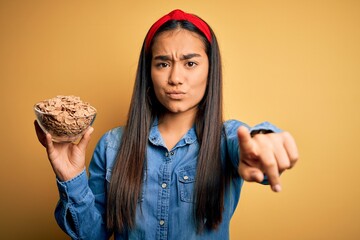 Canvas Print - Beautiful asian woman holding bowl with healthy corn flakes cereals over yellow background pointing with finger to the camera and to you, hand sign, positive and confident gesture from the front