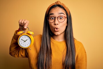 Wall Mural - Young asian woman holding vintage alarm clock standing over isolated yellow background scared in shock with a surprise face, afraid and excited with fear expression