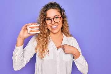 Wall Mural - Beautiful blonde woman with blue eyes holding plastic dentrure teeth over purple background with surprise face pointing finger to himself