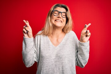 Poster - Young beautiful blonde woman wearing sweater and glasses over isolated red background gesturing finger crossed smiling with hope and eyes closed. Luck and superstitious concept.