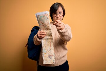 Sticker - Young down syndrome tourist woman wearing bag and looking at destination city map pointing with finger to the camera and to you, hand sign, positive and confident gesture from the front