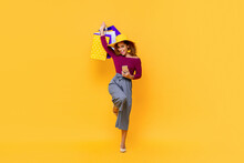 Fun Full Length Portrait Of Happy Young Attractive Shopaholic African American Woman Raising Shopping Bags And Holding Mobile Phone In Isolated Studio Yellow Background