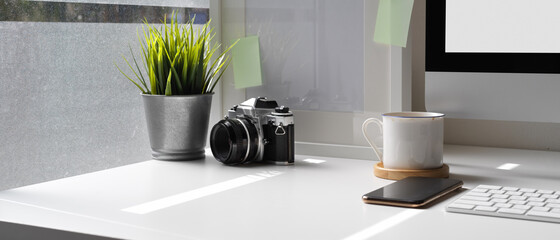 Modern office desk with computer device, smartphone, camera, supplies and decorations beside window