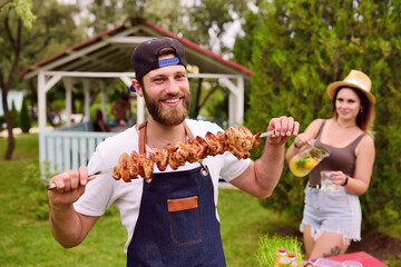 a young bearded man in a cap and denim apron smiles and holds a skewer with delicious barbecue meat cooked on the grill against the background of greenery, a Park and a gazebo.