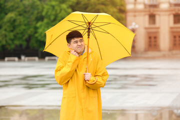 Wall Mural - Young Asian man in raincoat and with umbrella on city street