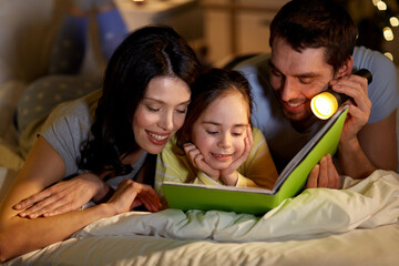 Poster - people and family concept - happy mother, father and little daughter reading book with torch light in bed at night at home