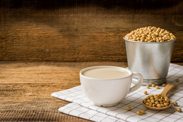 Soy milk in white cup healthy menu Designed with soybean seeds in bucket, Wooden spoon with white tablecloth on wooden floor.