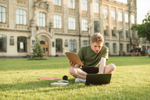 Concentrated Young Man In Casual Clothes Sits On Grass On Background Of A College Building And Studies With A Notebook In Hand And Laptop.Student On Campus Grass For A Break And Prepares For A Lesson