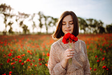Portrait Of A Beautiful Girl Having Fun Among The Red Flowers On The Poppy Field. Young Woman Smiling And Holding A Bouquet In Her Hands On The Sunset. Tender Romantic Moment Concept.