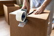 Close up man sealing cardboard box with adhesive tape, using dispenser, moving day and relocating delivery service concept, young male preparing to relocation, packing belongings, parcel