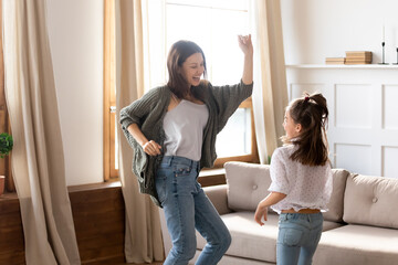 Happy excited mother and daughter dancing, listening to favorite song in modern living room, smiling mum and child having fun together, funny activity, moving to music, enjoying leisure time at home