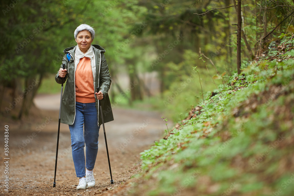 Full length portrait of active senior woman walking towards camera with Nordic poles while enjoying hike in beautiful autumn forest, copy space - obrazy, fototapety, plakaty 