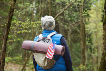 Wall Mural - back view at active senior man wearing backpack with american flag while enjoying hike in forest, co