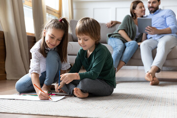 Children playing on warm floor while parents relaxing on couch, happy family spending weekend in living room, little sister and brother drawing with colorful pencils, playing together