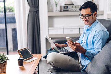 Wall Mural - Young smiling asian man relaxing using laptop computer working and video conference meeting at home.Young creative man looking at screen typing message with smartphone.work from home concept