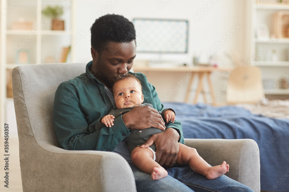 Portrait of caring African-American man holding cute mixed-race baby while sitting in comfortable armchair at home, copy space - obrazy, fototapety, plakaty 