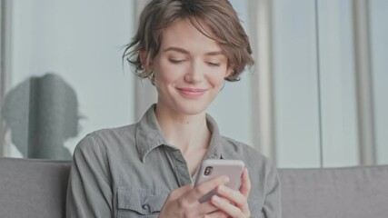 Poster - Happy young pretty woman using smartphone while sitting on sofa at home