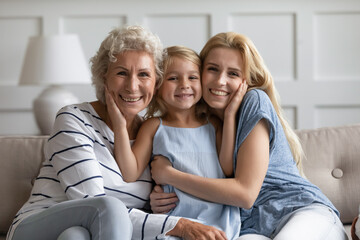 Three different age and generation relatives beautiful females sitting on sofa hugging posing looking at camera. Women or Mother Day holiday celebration, multigenerational family bond and ties concept