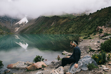 Wall Mural - Dreamy male tourist sitting on stone enjoying beauty of lake and amazing scenery of green mountains during wanderlust on holiday.Traveller resting during trip in exciting natural environment