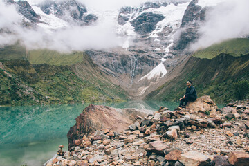 Wall Mural - Dreamy young man tourist in active wear admiring beauty of natural environment sitting on high hill for inspiration.Male traveller enjoying rest in mountains covered white clouds during hiking trek