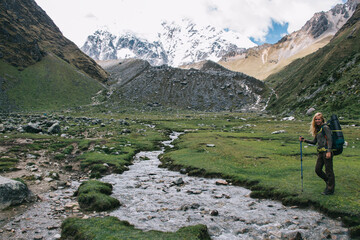 Poster - Portrait of positive young woman tourist with backpack and stick in hand smiling at camera while standing near river in Salkantay mountains.Happy female traveler with rucksack during hiking wanderlust