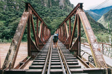 Poster - Back view of young woman tourist with backpack discover new places in wildness environment crossing bridge with rails over river.Hiker with travel rucksack going in mountains during trek