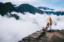 Woman With Blonde Hair Sitting In Lotus Pose On Edge Of Rock And Enjoying Calm And Inspiration For Found Soul Harmony During Travel Tour To Mountain And Machu Picchu In Peru Covered With Clouds