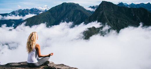 Wall Mural - Back view of woman tourist sitting in lotus pose relaxing and admiring breathtaking view during trek tour, female traveler enjoying beauty of Machu Picchu landscape and high mountains covered clouds