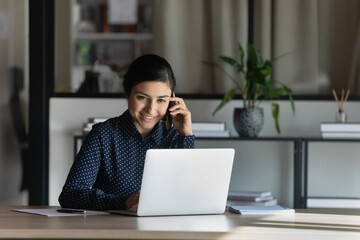 Canvas Print - Smiling Indian businesswoman feels satisfied confident provide help consulting client distantly by mobile phone call seated at desk in modern office. Busy fruitful workday, successful employee concept