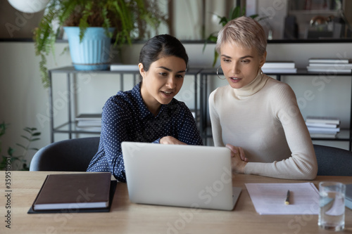 Multiethnic girls colleagues indian and caucasian young women working together sit at workplace desk using laptop discussing app, mentor teach apprentice feels confident provide help to intern concept