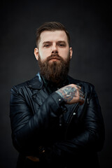 Wall Mural - Cool bearded tattooed man in a leather jacket looking on the camera in a dark studio