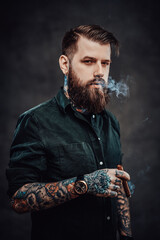 Wall Mural - Stylish bearded man smokes a cigar. Studio portrait of a handsome tattooed hipster guy who smokes a cigar