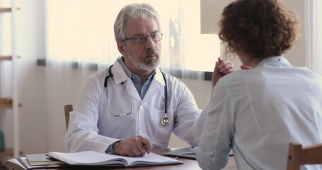 Wall Mural - Serious focused middle aged hoary physician in medical coat sitting at table, consulting female patient about illness or surgery. Rear view young woman listening to old doctor at checkup meeting.