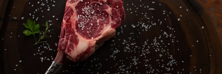 Wall Mural - Top view raw tomahawk beef steak with ingredients for grilling with a spice rub, fresh rosemary and salt , on a table with dark wood texture and space for writing