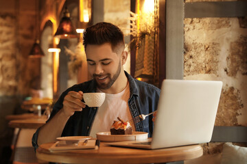 Wall Mural - Young blogger with laptop eating dessert and drinking coffee at table in cafe