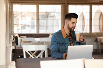 Wall Mural - Young blogger with laptop drinking latte at table in cafe. Space for text