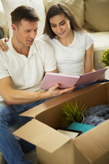 Cute couple unpacking cardboard boxes in their new home, sitting on the floor and looking at a family album