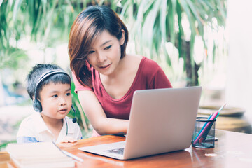Wall Mural - Young asian mother and son using laptop computer for study and learning together at home, boy wearing headphone for e-learning with distancing, teacher or mom support child, education concept.