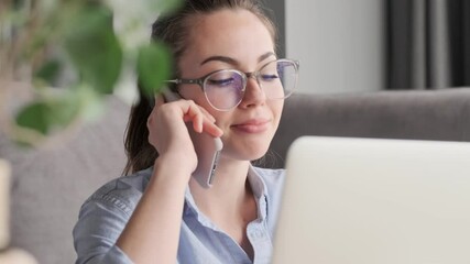 Wall Mural - Smiling attractive brunette woman in eyeglasses talking by smartphone while sitting with laptop computer on the floor near sofa at home