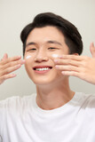 Fototapeta Do pokoju - Close-up portrait of a smiling, happy-looking young asian man with moisturizer cream applied on the face.