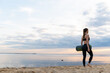 © kalinichenkod - young fitness model with yoga mat leaves at sunset against the sky
