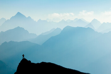 Poster - Man reaching summit after climbing and hiking enjoying freedom and looking towards mountains silhouettes panorama in early Morning.