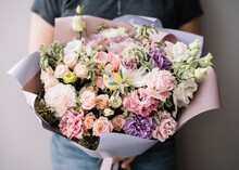 Very Nice Young Woman Holding Big And Beautiful Blossoming Bouquet Of Fresh Eustoma, Roses, Carnations Flowers In Pastel Purple And Peachy Colors On The Grey Wall Background 
