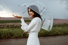 Young Woman Holding A Clear, Transparent Umbrella On A Stormy Weather, Standing In An Open Field, Checking The Rain.